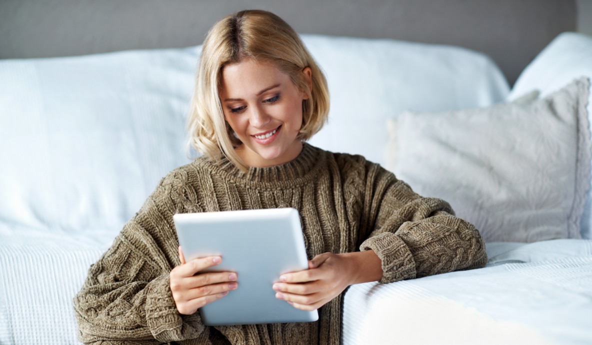 Mulher sentada em um sofá usando um tablet com uma estante ao fundo.