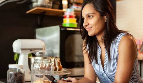 Mulher de blusa listrada, encostada na bancada da cozinha, no celular