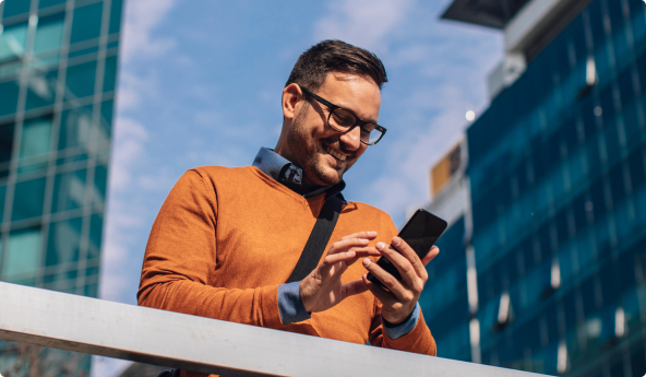 Homem com blusa laranja e óculos, mexendo no celular, ao ar livre