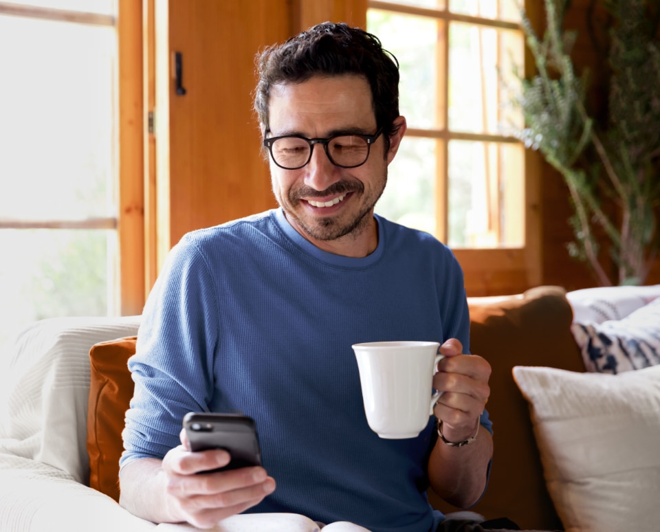 Fotografia de homem de meia idade, de óculos de grau e blusa azul, sorridente, recebendo mensagem no seu celular, com uma caneca de café em sua casa.