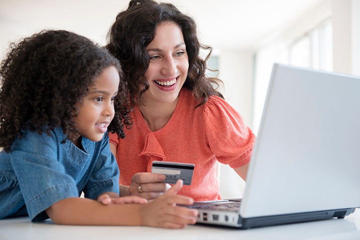 Mulher segurando o cartão Itaú junto com uma criança e usando o notebook representando a compra parcelada com juros 