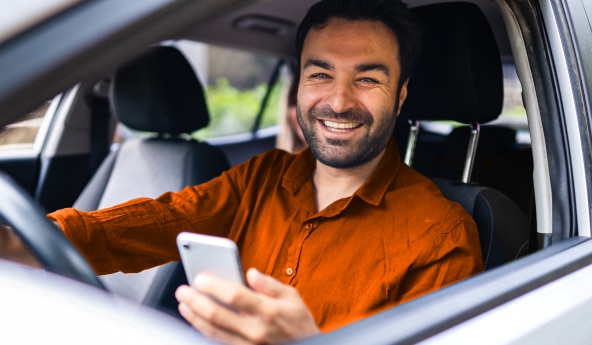 Homem sorrindo com um celular na mão enquanto está parado com seu carro