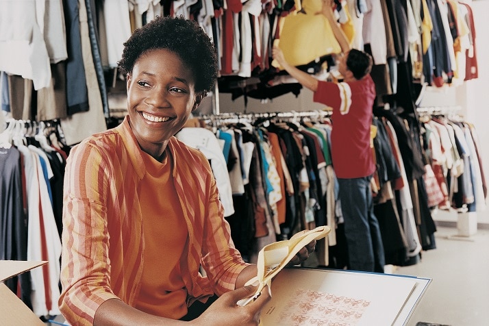 Mulher sorrindo na sua loja de roupas, enquanto um funcionário está ajustando os cabides atrás dela