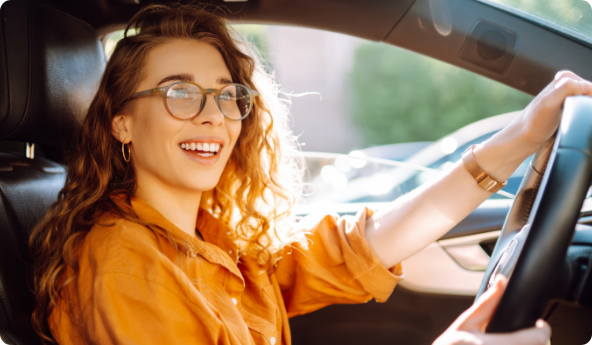 Mulher de óculos e camisa laranja, sorrindo, enquanto está com as mãos no volante de seu carro